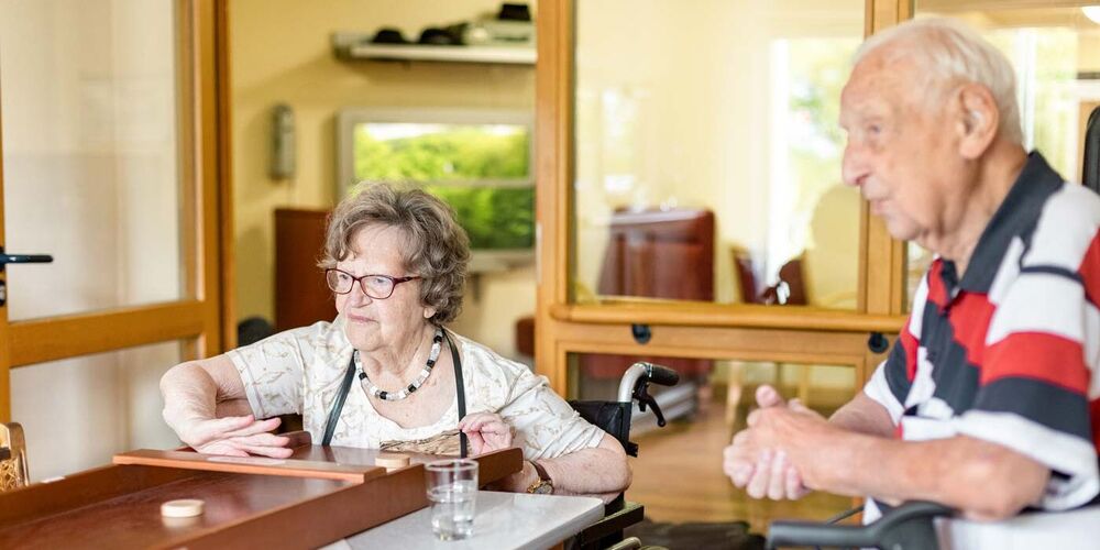 Ein Senior und eine Seniorin in einem Rollstuhl sitzen vor einem Shuffleboard und spielen an diesem.
