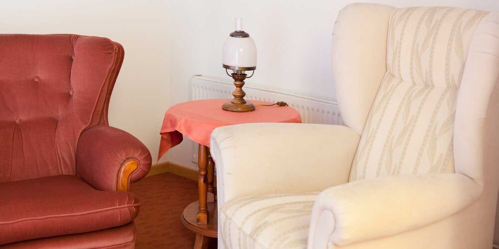 Ein Sessel und ein Sofa zwischen welchen ein kleiner Tisch mit einer Lampe steht. 